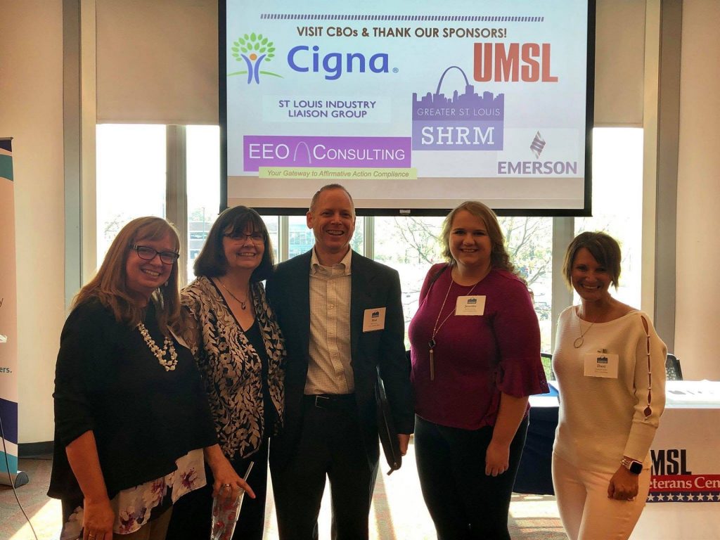 Five people wearing name badges standing in front of a screen thanking the event sponsors, EEO Consulting among them, at a St. Louis Industry Liaison Group event