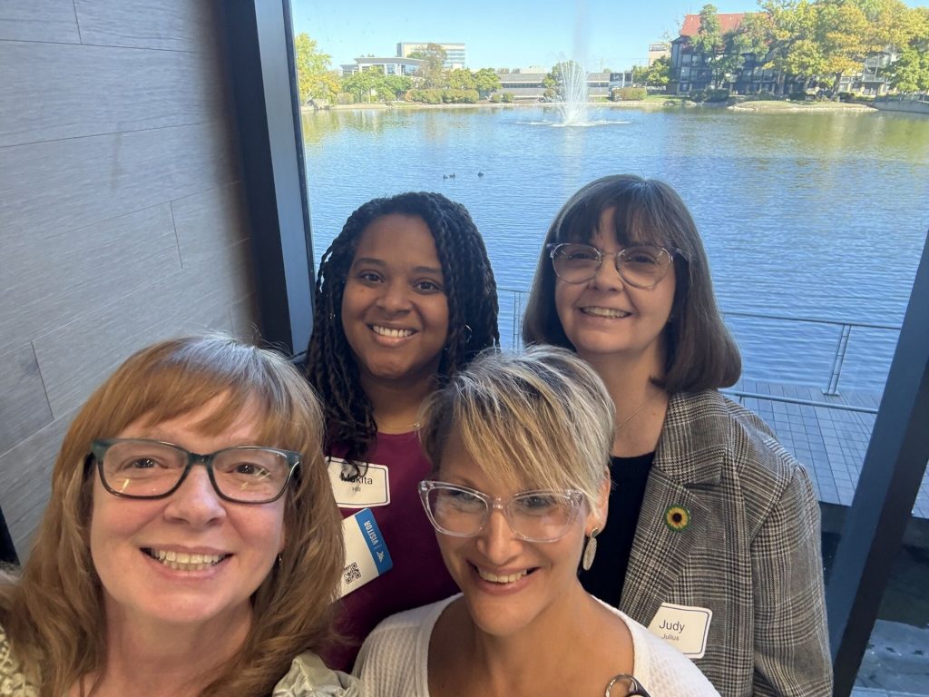 Four people wearing conference name badges, including Judy Julius, smiling together beside a window overlooking a lake and fountain